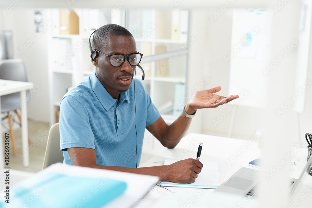 African young man in casual clothing sitting at the table wearing headphones and explaining something on the phone he working in a call center