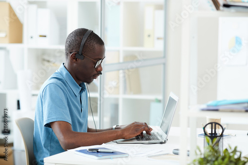 Serious African programmer in casual clothing wearing headphones and concentrating on his work on laptop computer at office