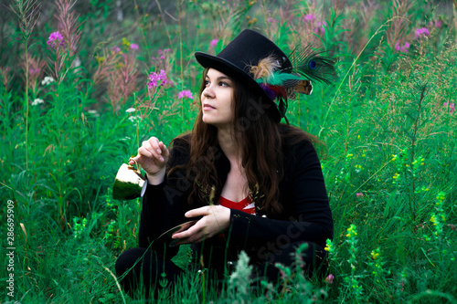 A woman in a tall hat sits in the grass with a mug in her hands
