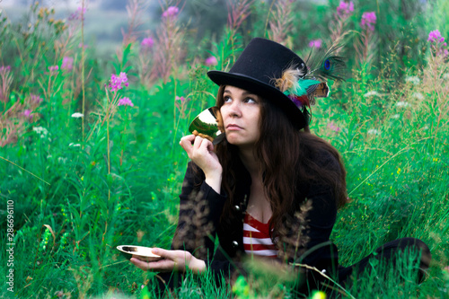 A woman in a tall hat sits in the grass with a mug in her hands