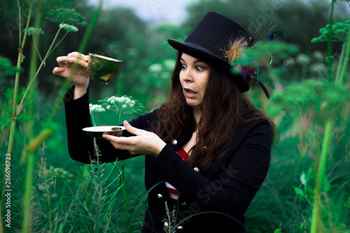 A woman in a tall hat in a clearing with purple and white tall flowers with a mug in her hands