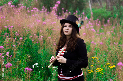A woman in a tall hat in a clearing with purple flowers with a recorder in her hands