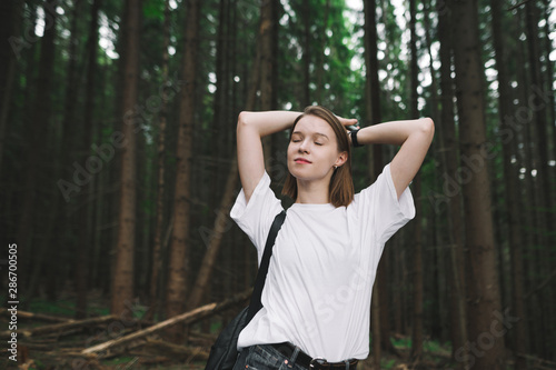 Young attractive female traveler standing in the mountain woods, hands up and behind her head, eyes closed, feeling satisfied and relaxed. Portrait of a dreamy tourist girl in the mountain fir forest.