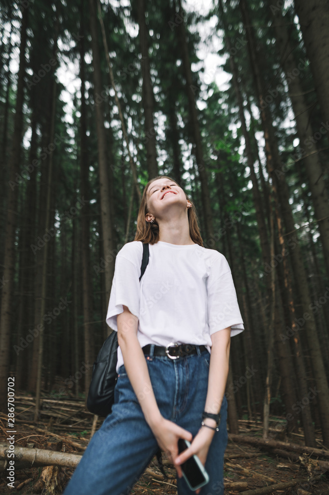 Vertical photo of smiling tourist girl with her eyes closed feeling happy walking in the coniferous forest. Young caucasian female traveler with broad smile being positive and cheerful in the woods. Vertical photo of smiling tourist girl with her eyes closed feeling happy walking in the coniferous forest. Young caucasian female traveler with broad smile being positive and cheerful in the woods.