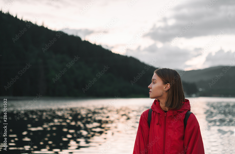 Traveler girl looking aside relaxing near the mountain forest and lake, enjoying beautiful view in the evening. Calm portrait of a young woman in a red raincoat near the mountain pond. Copy space. Traveler girl looking aside relaxing near the mountain forest and lake, enjoying beautiful view in the evening. Calm portrait of a young woman in a red raincoat near the mountain pond. Copy space.
