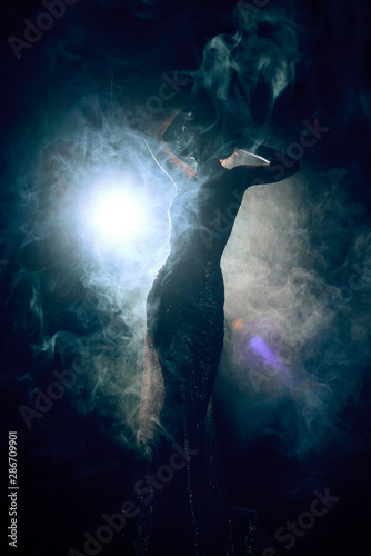 Silhouette of a slim, artistic girl wearing an elegant evening dress in theatrical fog. Sensual, conceptual and fashionable design. Copy space