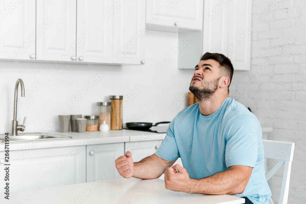 angry man gesturing while sitting near table in kitchen angry man gesturing while sitting near table in kitchen
