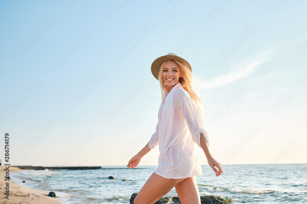 Young gorgeous smiling blond woman in shirt and hat happily walking by the sea