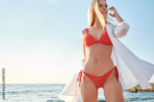 Young beautiful blond woman in swimsuit and white shirt dreamily looking aside on beach with sea on background