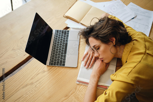 Top view of young tired woman dreamy asleep on desk with laptop and documents under head at workplace photo