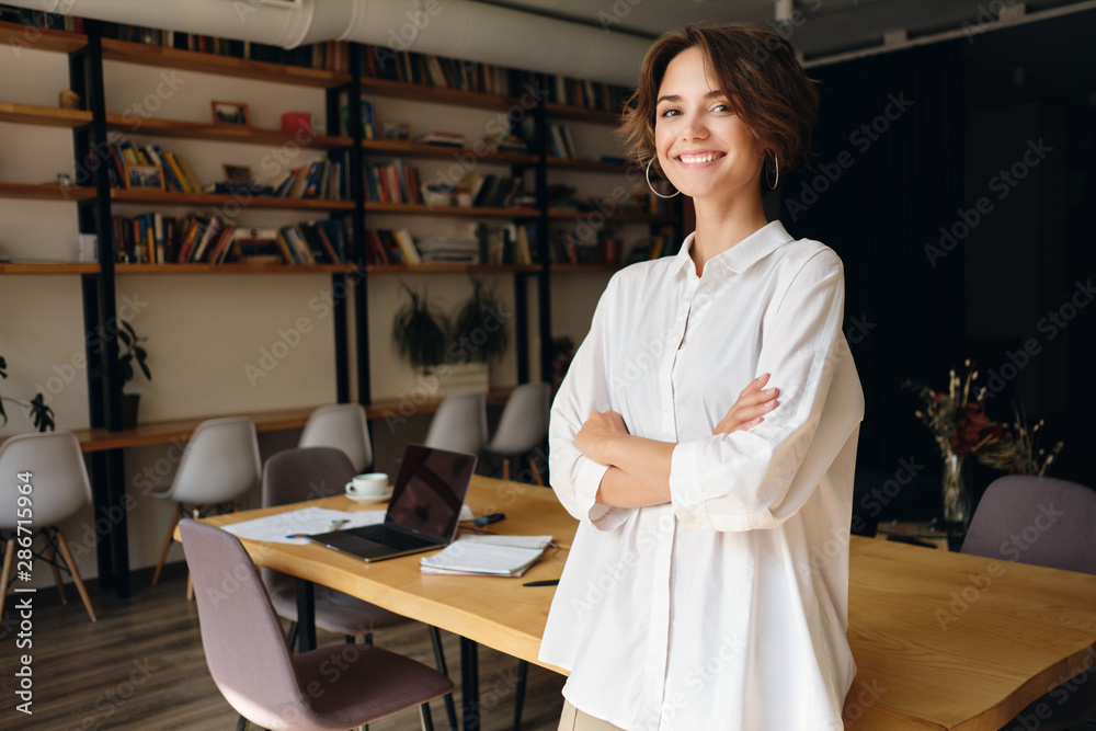 Young cheerful woman in white shirt happily looking in camera with desk on background in office - obrazy, fototapety, plakaty 