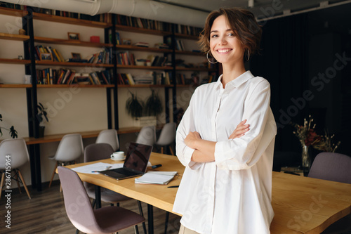 Young cheerful woman in white shirt happily looking in camera with desk on background in office photo