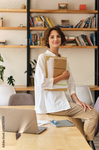 Young attractive smiling woman in white shirt happily looking in camera sitting on desk with papers and laptop in office