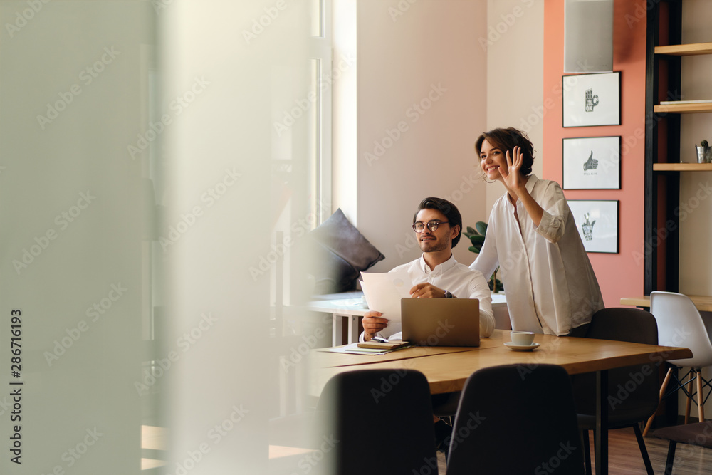 Young smiling business colleagues with papers happily greeting someone at work in office