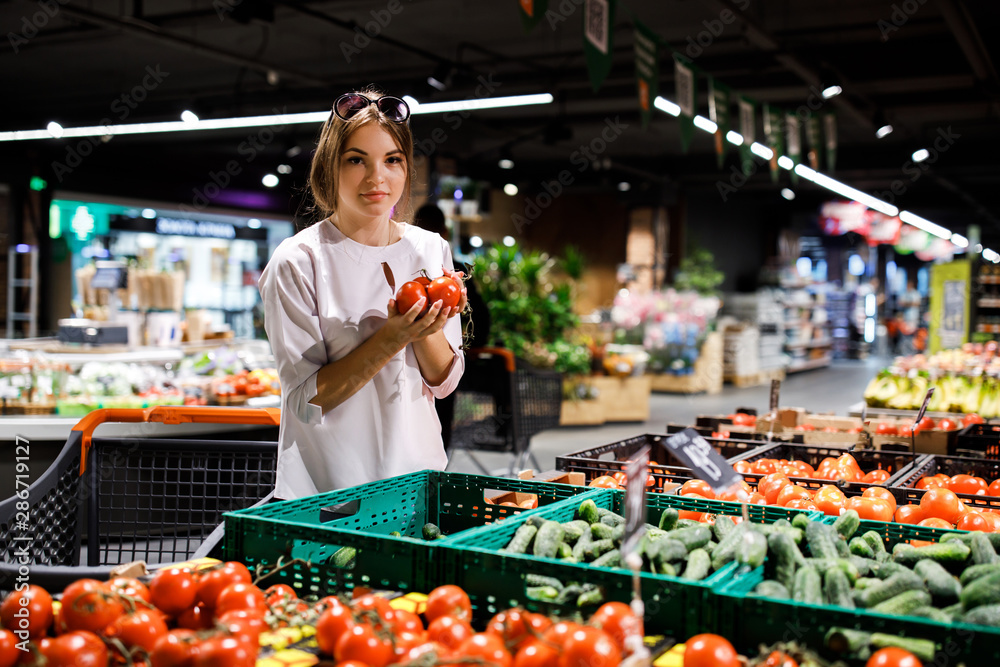 Obraz premium Young pretty girl is shopping in a big store. The girl buys groceries at the supermarket. Girl chooses fresh vegetables