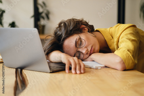 Young tired woman in eyeglasses sleeping on desk with laptop at workplace alone photo