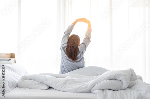 An Asian woman wakes up in the morning on the white bed of a hotel room with stretching.