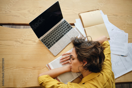 Top view of young tired woman asleep on desk with laptop and documents under head at workplace