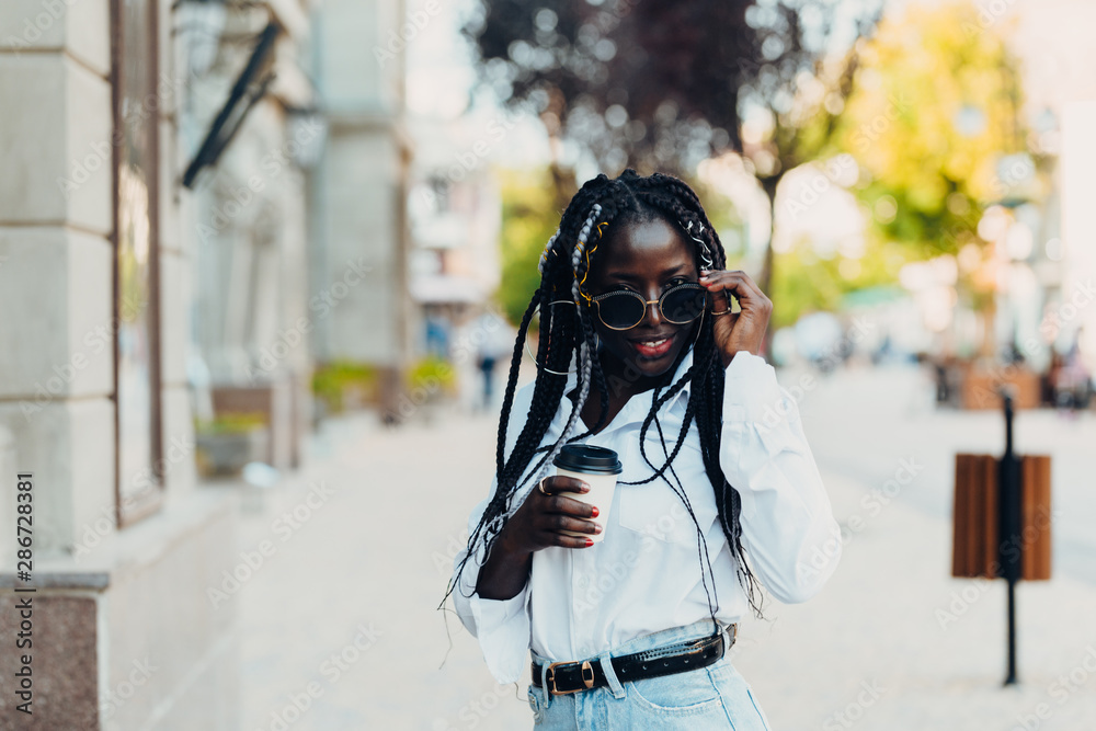Portrait of a smiling young dark-skinned girl with pigtails in glasses with coffee in hand outdoors on a sunny day. Portrait of a smiling young dark-skinned girl with pigtails in glasses with coffee in hand outdoors on a sunny day.