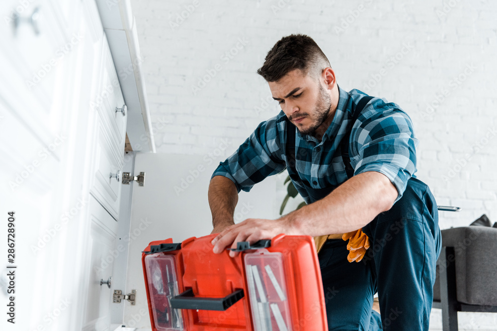 selective focus of handsome handyman looking at opened toolbox in kitchen selective focus of handsome handyman looking at opened toolbox in kitchen