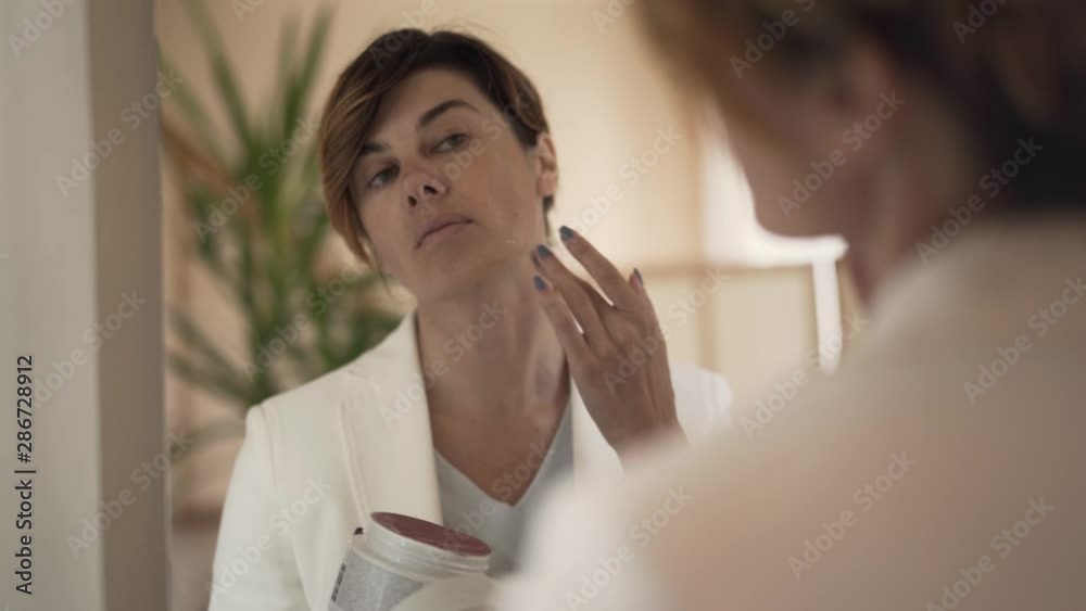 Portrait of an elegant well-dressed lady applying a cream on her face looking in the mirror. The woman preparing for the work in the morning. Wellness, body care, face care Portrait of an elegant well-dressed lady applying a cream on her face looking in the mirror. The woman preparing for the work in the morning. Wellness, body care, face care
