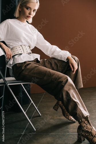 stylish blonde woman in white blouse and boots with snakeskin print sitting on chair near curtain on brown