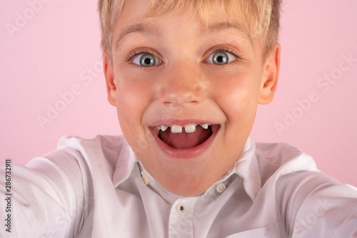 Cheerful selfie. Closeup of cheerful young boy in shirt holding mobile phone and making photo of himself while standing against pink background