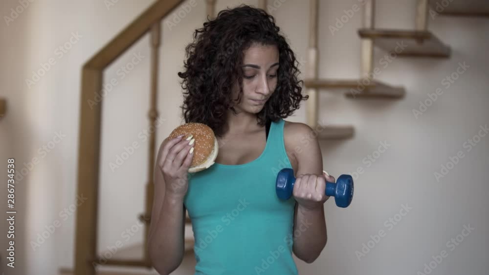 Portrait of an attractive slim lady holding a big burger and a small dumbbell in hands trying to choose between a healthy lifestyle and tasty food. Choice, dilemma concept. Portrait of an attractive slim lady holding a big burger and a small dumbbell in hands trying to choose between a healthy lifestyle and tasty food. Choice, dilemma concept.