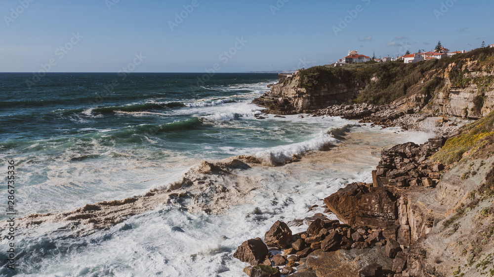 View of cliff top villas and the Atlantic ocean waves on a sunny summer day, taken in Praia das Maçãs, Sintra, Portugal