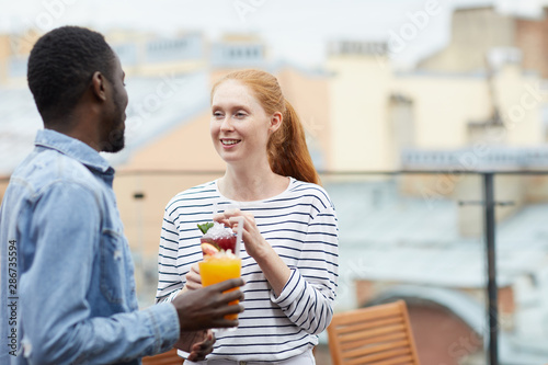 Young red haired woman talking to young African man and smiling while they standing and drinking fresh juice together