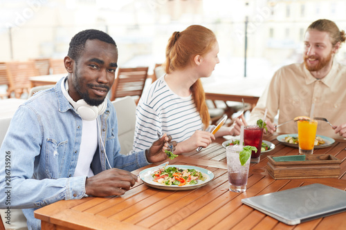 African young man eating vegetable salad and smiling at camera while sitting at the table with his friends during lunch in cafe