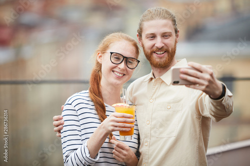Young man embarcing young woman with cocktail and they smiling at camera while making selfie portrait on mobile phone outdoors