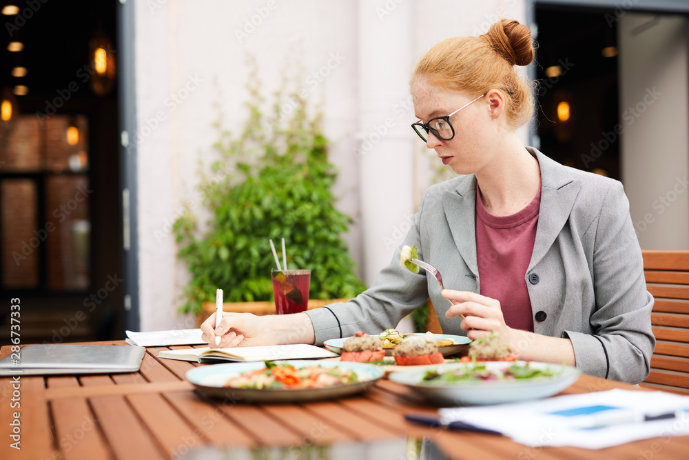 Serious businesswoman in eyeglasses making notes in her notepad while sitting at the table and eating vegetable salad during lunch in cafe Serious businesswoman in eyeglasses making notes in her notepad while sitting at the table and eating vegetable salad during lunch in cafe
