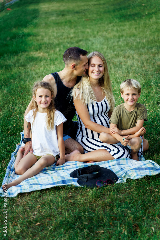Picture of kissing parents with children sitting on plaid on picnic on summer day