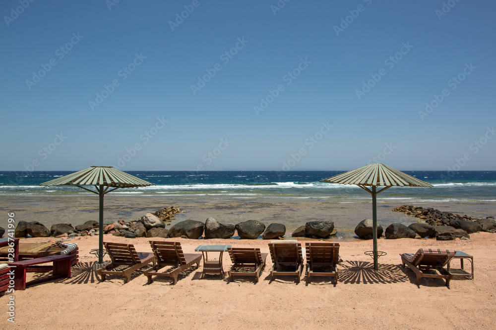 Wooden sun loungers and parasols on the sandy beach of the Red Sea. Place to relax and spend a vacation. The wave goes to the shore. Dahab, Egypt Wooden sun loungers and parasols on the sandy beach of the Red Sea. Place to relax and spend a vacation. The wave goes to the shore. Dahab, Egypt