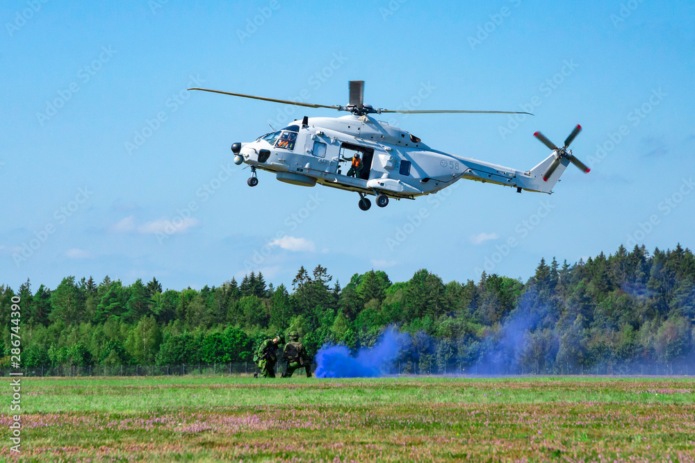 Landing of swedish military helicopter NH90 HKP 14. Military armed men with blue smoke bomb near forest under blue sky Landing of swedish military helicopter NH90 HKP 14. Military armed men with blue smoke bomb near forest under blue sky