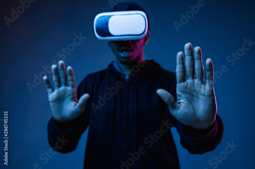 Young african-american man's playing in VR-glasses in neon light on gradient background. Male portrait. Concept of human emotions, facial expression, modern gadgets and technologies. Excited, shocked.