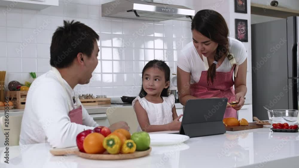 Happy Asian family make a cooking. Father, Mother and Daughter are preparing fruit orange and see Digital tablet in the kitchen at home. Healthy food concept and happy holidays Happy Asian family make a cooking. Father, Mother and Daughter are preparing fruit orange and see Digital tablet in the kitchen at home. Healthy food concept and happy holidays