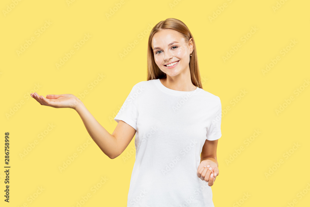 Caucasian young woman's half-length portrait on yellow studio background. Beautiful female model in white shirt. Concept of human emotions, facial expression. Showing and pointing something. Caucasian young woman's half-length portrait on yellow studio background. Beautiful female model in white shirt. Concept of human emotions, facial expression. Showing and pointing something.
