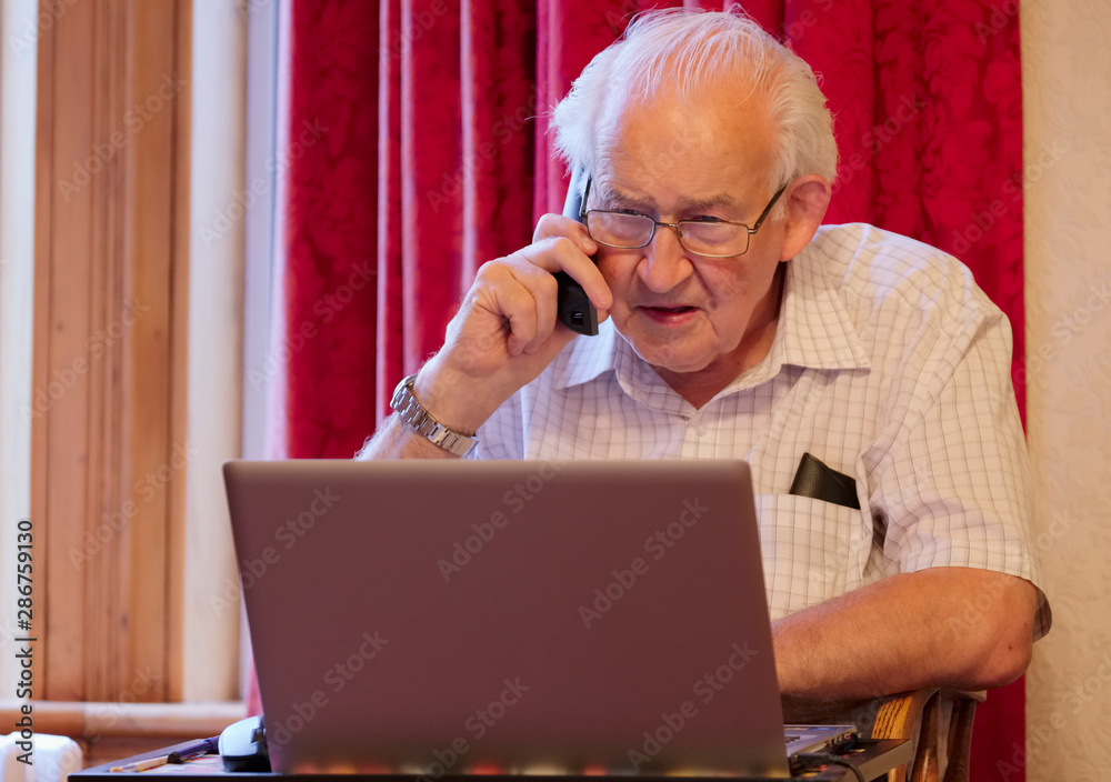 Old elderly senior man on phone at laptop computer at risk to cyber attack and online bank fraud