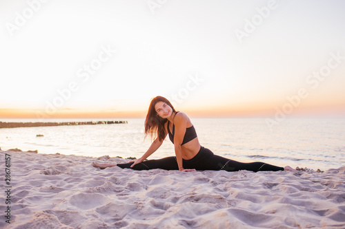Slim strong young woman in black practicing yoga doing the splits on sand beach close-up with copy space. Sunset, summer. Healhy lifestyle concept.