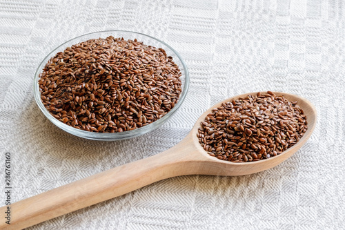 Glass bowl and wooden spoon are full of whole flax seeds on a linen cloth background. Healthy food for lower cholesterol and preventing heart diseases.