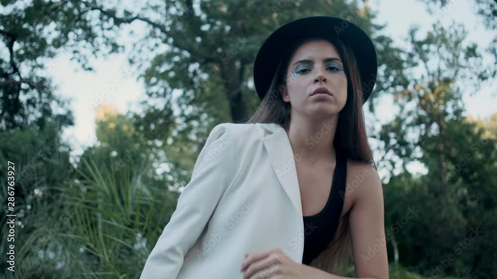 Tracking out shot of beautiful fashion girl in hat and jacket posing confidently looking in camera in old garden Tracking out shot of beautiful fashion girl in hat and jacket posing confidently looking in camera in old garden