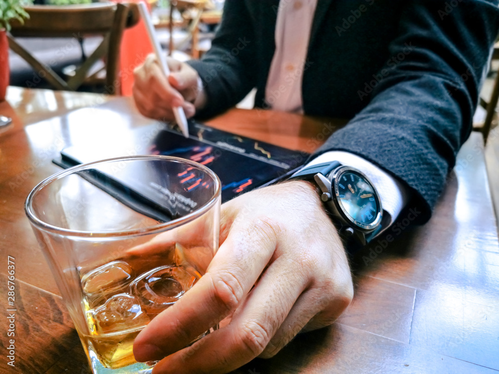 business man with beard who has smart watch using pro tablet and pencil interacting with tablet screen holographic economy icons drinking whiskey in the cafe pub