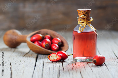 Photography Glass bottle of rosehip seed essential oil with fresh rose hip fruits in shovel on wooden rustic background