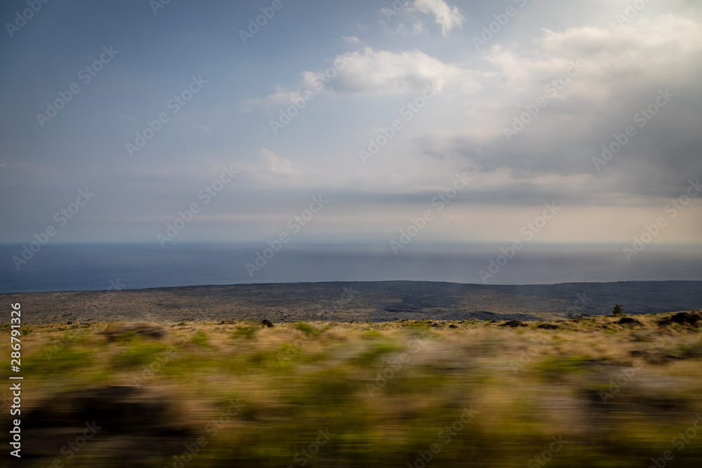 The foreground whizzes by looking out to a flat ocean fading into the horizon with a dark lava field and dry yellow and green bushes from a moving vehicle in Hawaii