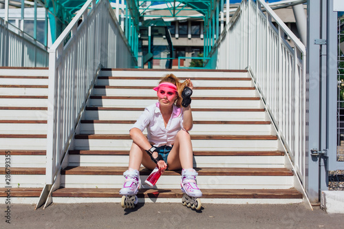 Portrait of an emotional girl in a pink cap visor wearing protective gloves and rollerblades sitting on stairs holding a bottle of water in hands photo