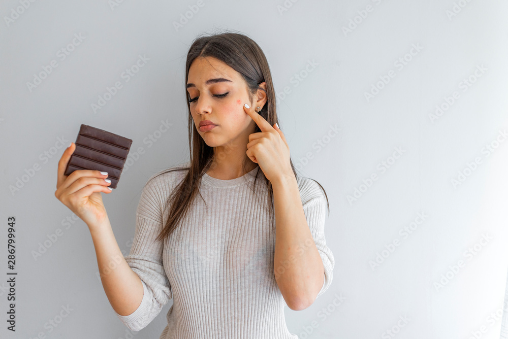 Young woman with acne problem holding chocolate bar on grey background