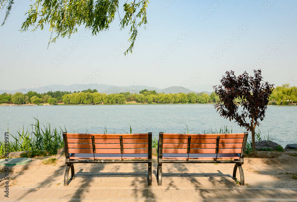 Two empty benches facing the lake with a background of trees and mountains in Yuanmingyuan Ruins Park, Beijing, China.
