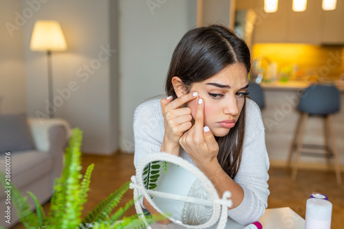 Portrait of beautiful young woman squeezing pimples while looking at the mirror. Pimple on cheek. Young woman squeeze her acne in front of the mirror. I'm sure this will go away if I pop it. photo